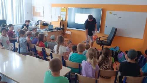 Uczniowie siedzą przed tablicą multimedialną, nauczyciel siedzi przy biurku. Gość podaje dzieciom rzeczy, jakie można drukować 3D.